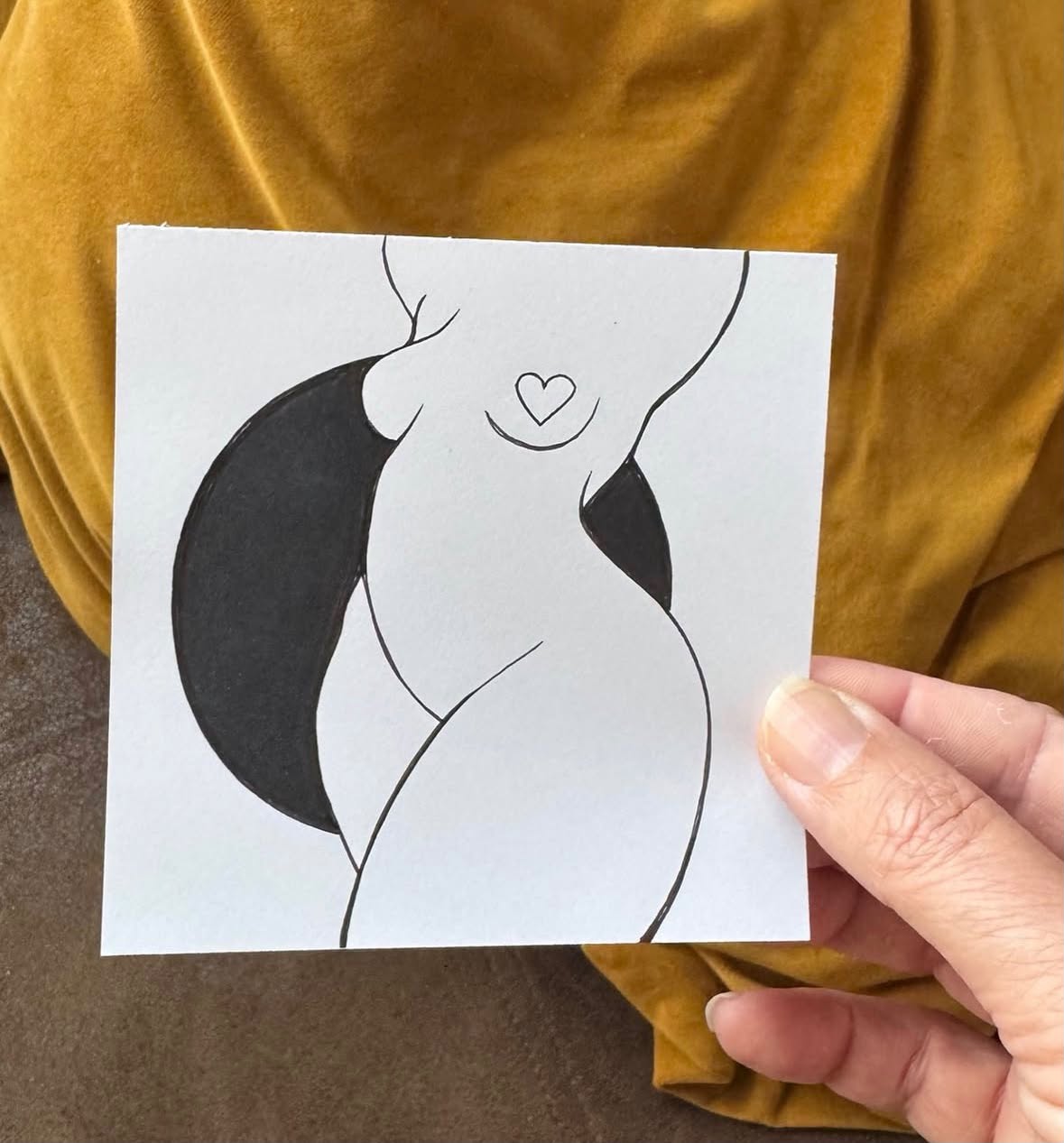 A hand holds a black-and-white Being drawing by Hanneke Wetzer against an ochre-yellow background.